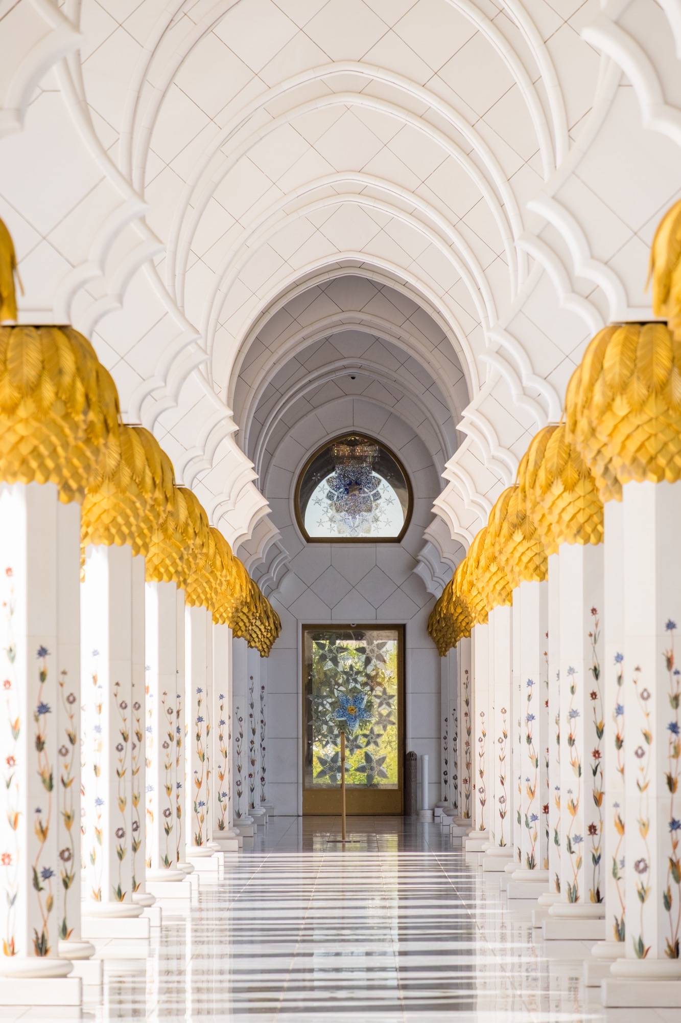 Wnętrze meczetu z białymi kolumnami zdobionymi kwiatami i złotymi detalami, perspektywa prowadząca do okna z witrażem.