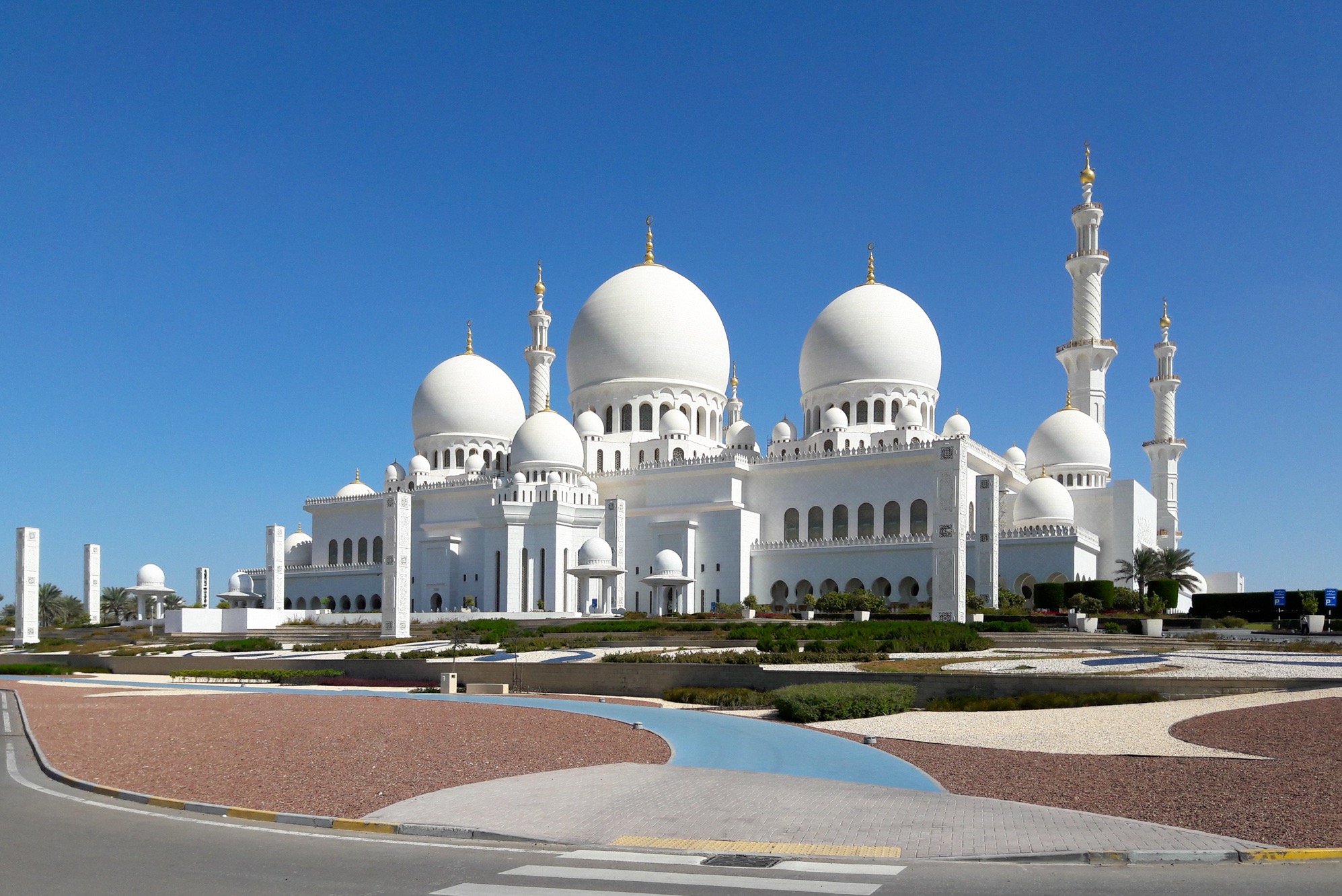 Imponujący widok na biały meczet z kopułami i minaretami na tle błękitnego nieba. Architektura islamska, detale konstrukcyjne, starannie zaprojektowany krajobraz.