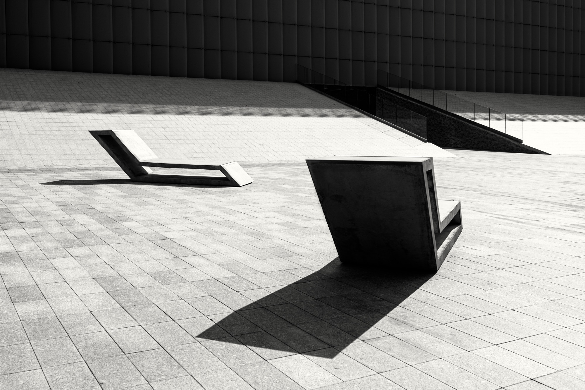 Minimalistyczna architektura miejska w czerni i bieli: betonowe ławki, geometryczne wzory kostki brukowej i fasada budynku w tle, oświetlone mocnym światłem słonecznym.