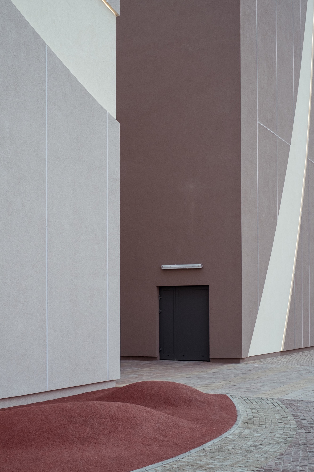 Minimalistyczna architektura budynku z elementami w odcieniach szarości i brązu, z akcentem na geometryczne linie i faktury ścian oraz nietypowe ukształtowanie terenu.