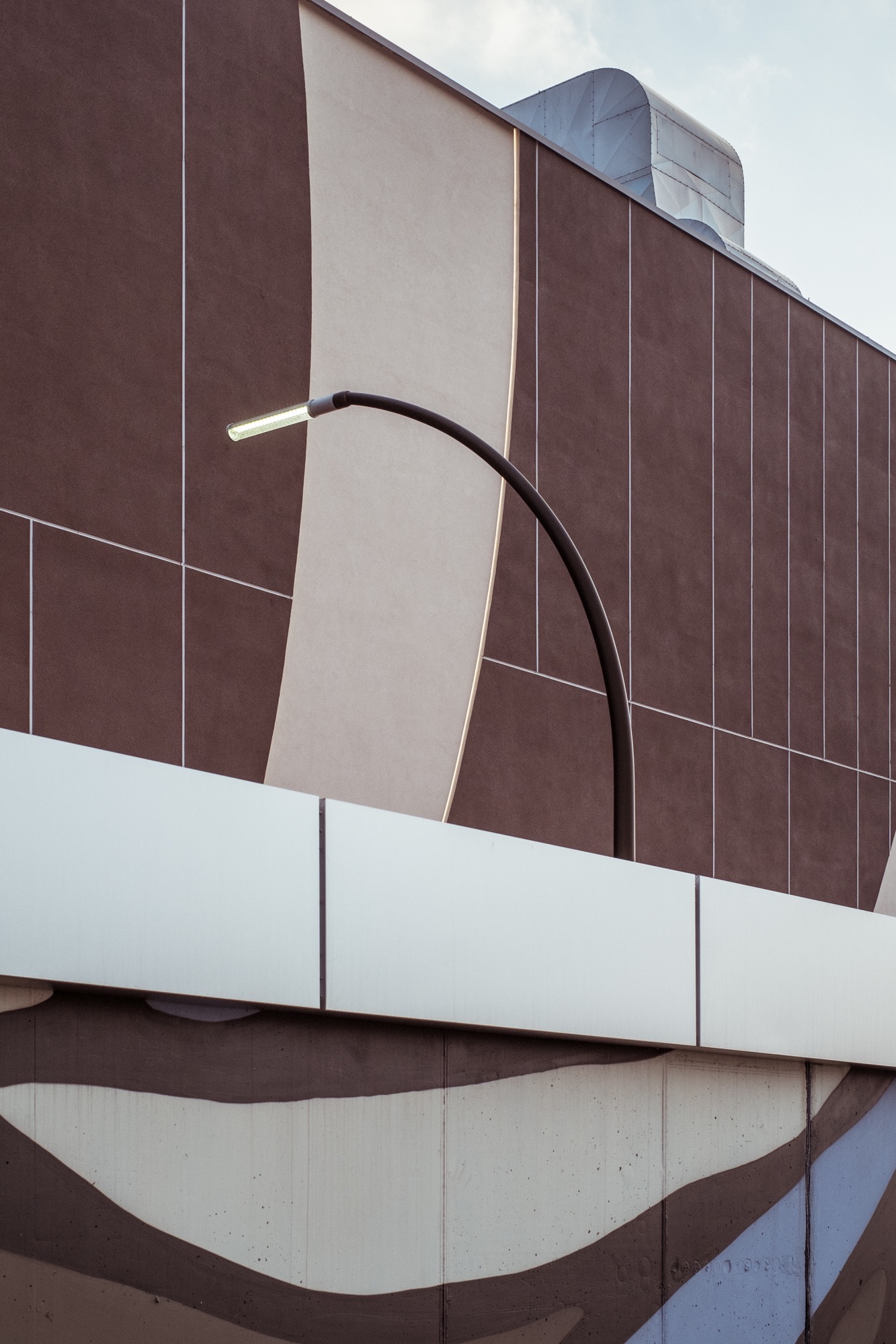 Fasada budynku z geometrycznym wzorem w odcieniach brązu i beżu, oświetlona nowoczesną lampą uliczną. Widoczny fragment instalacji wentylacyjnej na dachu.