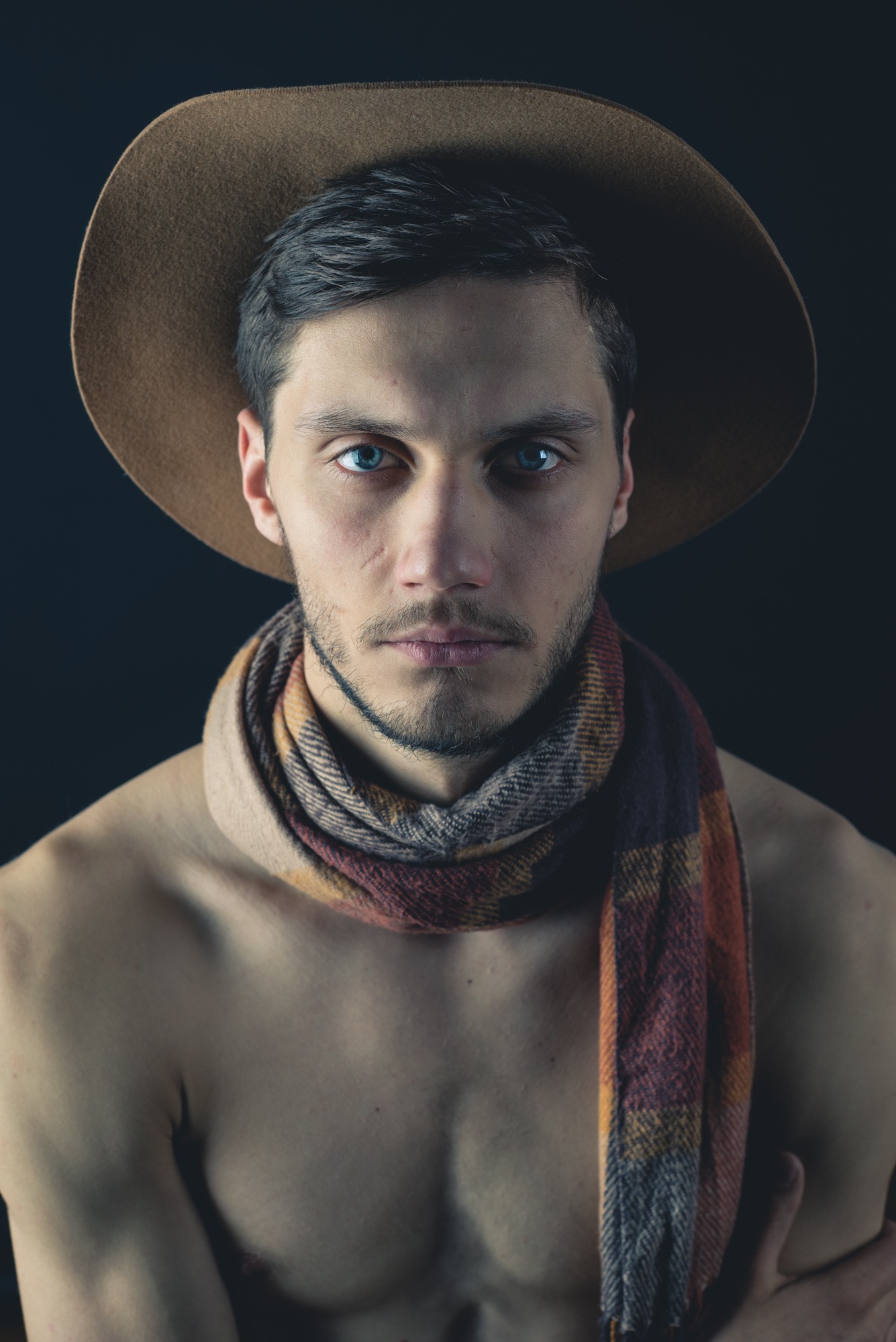 Portret mężczyzny w kapeluszu i szaliku na nagim ciele, z intensywnym spojrzeniem i ciemnym tłem, ukazujący artystyczny styl i dbałość o detale.