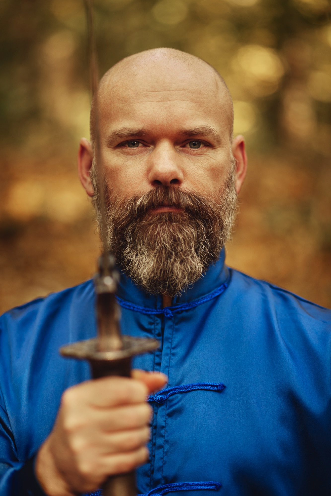 Portret mężczyzny z brodą w niebieskim stroju, trzymającego miecz na tle jesiennego lasu. Ujęcie z bliska, ostre światło podkreśla detale twarzy.