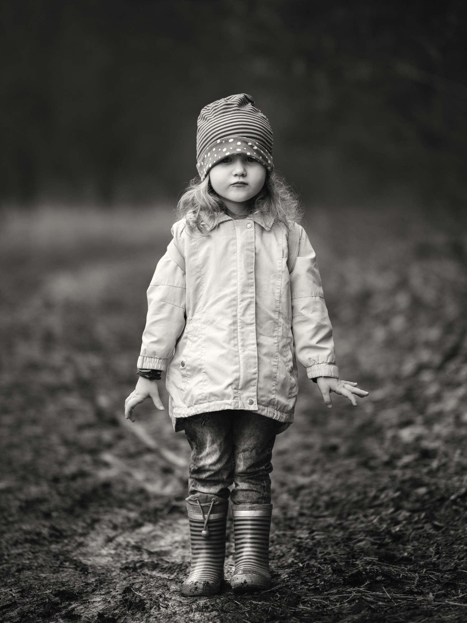 Monochromatyczny portret dziewczynki w paski czapce i kaloszach, stojącej na błotnistej ścieżce. Uwagę przykuwa jej poważny wyraz twarzy i naturalne otoczenie.