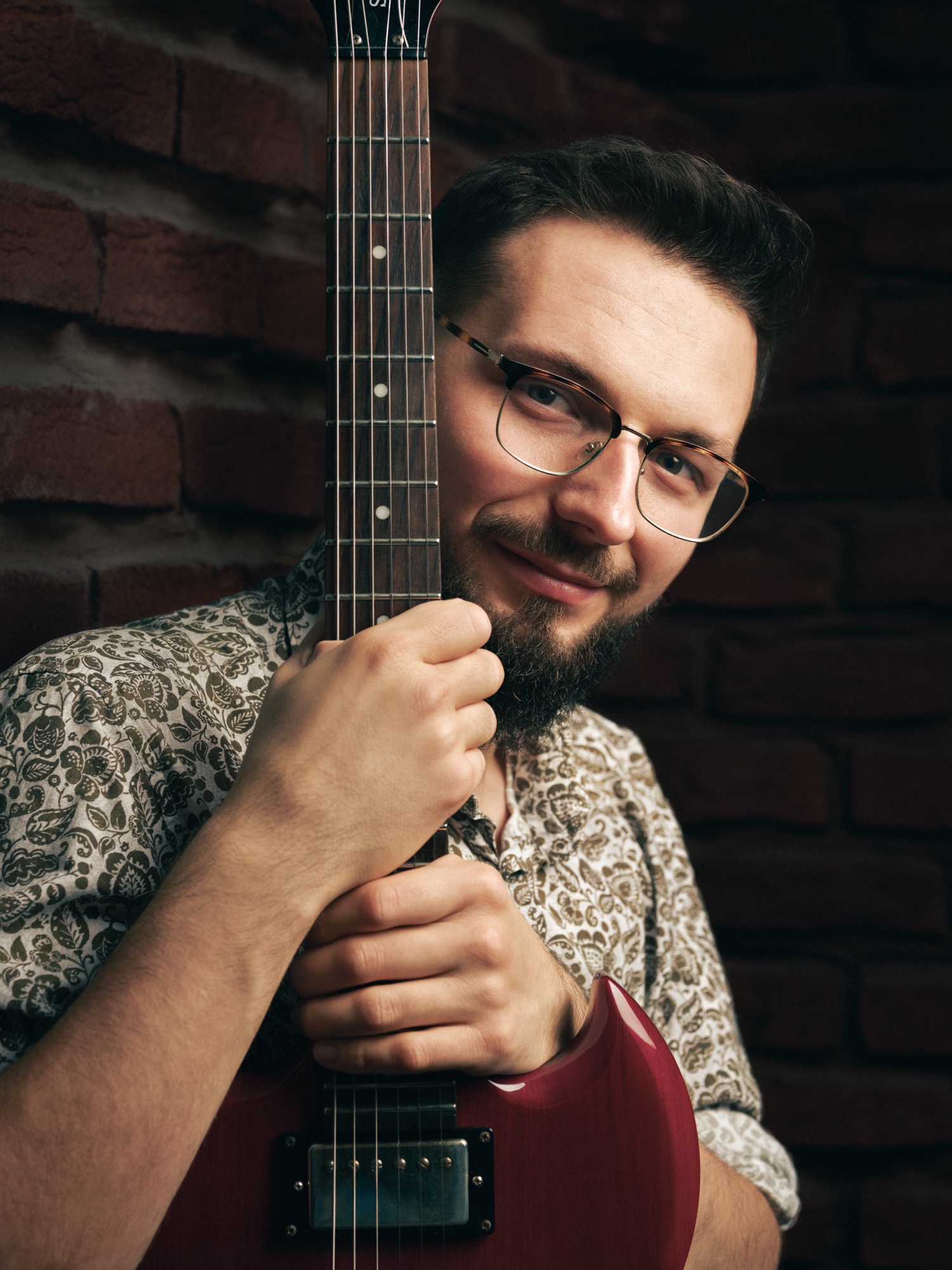 Uśmiechnięty mężczyzna w okularach, opierający się o czerwoną gitarę na tle ceglanej ściany. Portret muzyka z instrumentem w studiu.