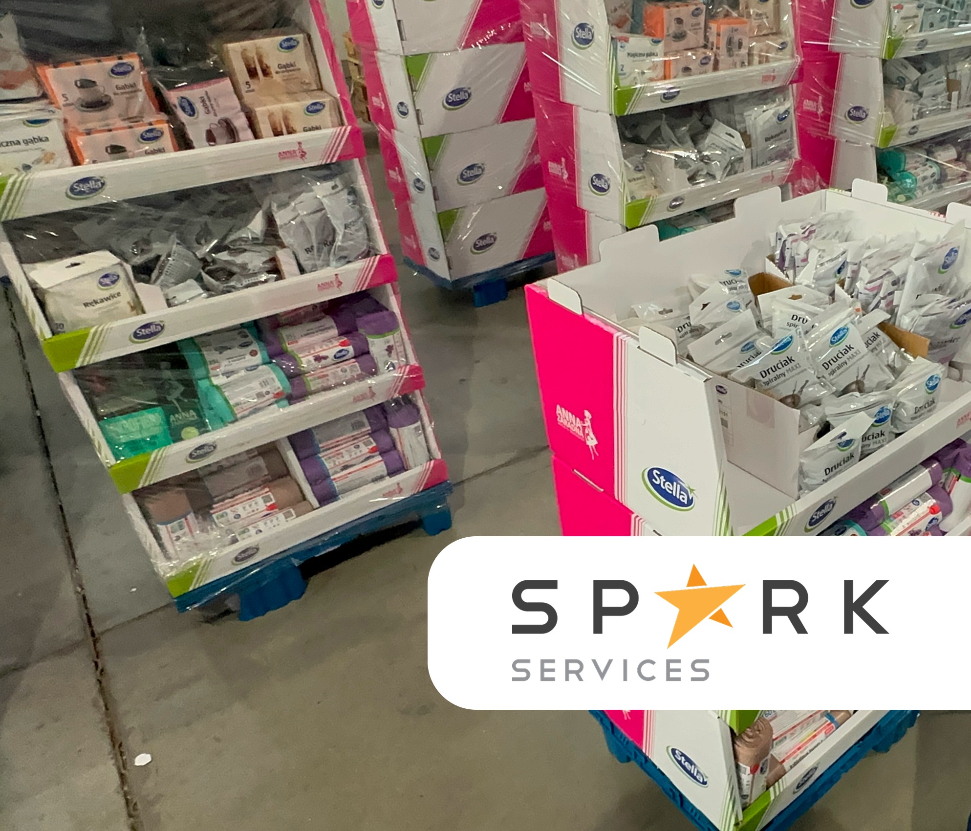 Ekspozytory kartonowe z produktami marki Stella na paletach, owinięte folią. Różne rodzaje produktów w opakowaniach, gotowe do transportu i sprzedaży.