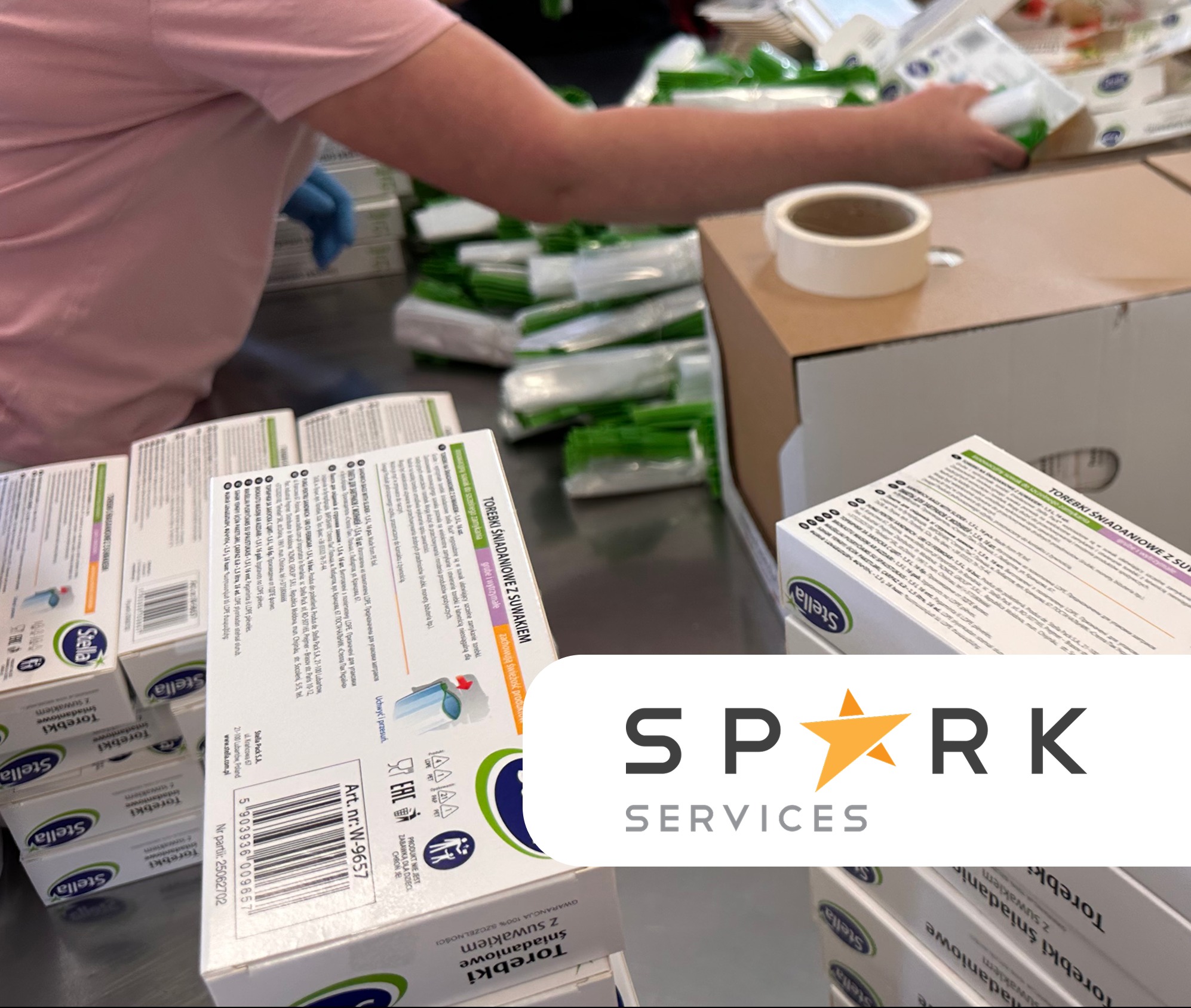 Pakowanie torebek śniadaniowych z suwakiem w kartony. Widoczne opakowania jednostkowe i rolka taśmy klejącej. Praca wykonywana w rękawicach ochronnych.