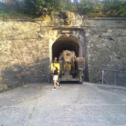 Firma Handlowo-Usługowa Piotr Czyszczoń - Wnętrze Twierdzy Kłodzkiej z maszyną górniczą i pracownikami. Kamienne mury, tunel, oświetlenie. Osoba w krótkich spodenkach idzie w kierunku maszyny.