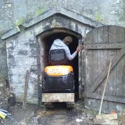 Firma Handlowo-Usługowa Piotr Czyszczoń - Mini koparka Kubota w kamiennym wejściu z drewnianymi drzwiami, prawdopodobnie w Twierdzy Kłodzko. Osoba wewnątrz. Prace ziemne w toku.