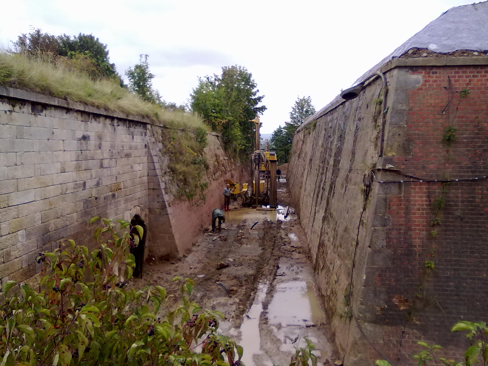 Prace ziemne w wąskim wykopie, prawdopodobnie pod instalację pionowych wymienników ciepła. Widoczna wiertnica i pracownicy w trudnych warunkach terenowych.