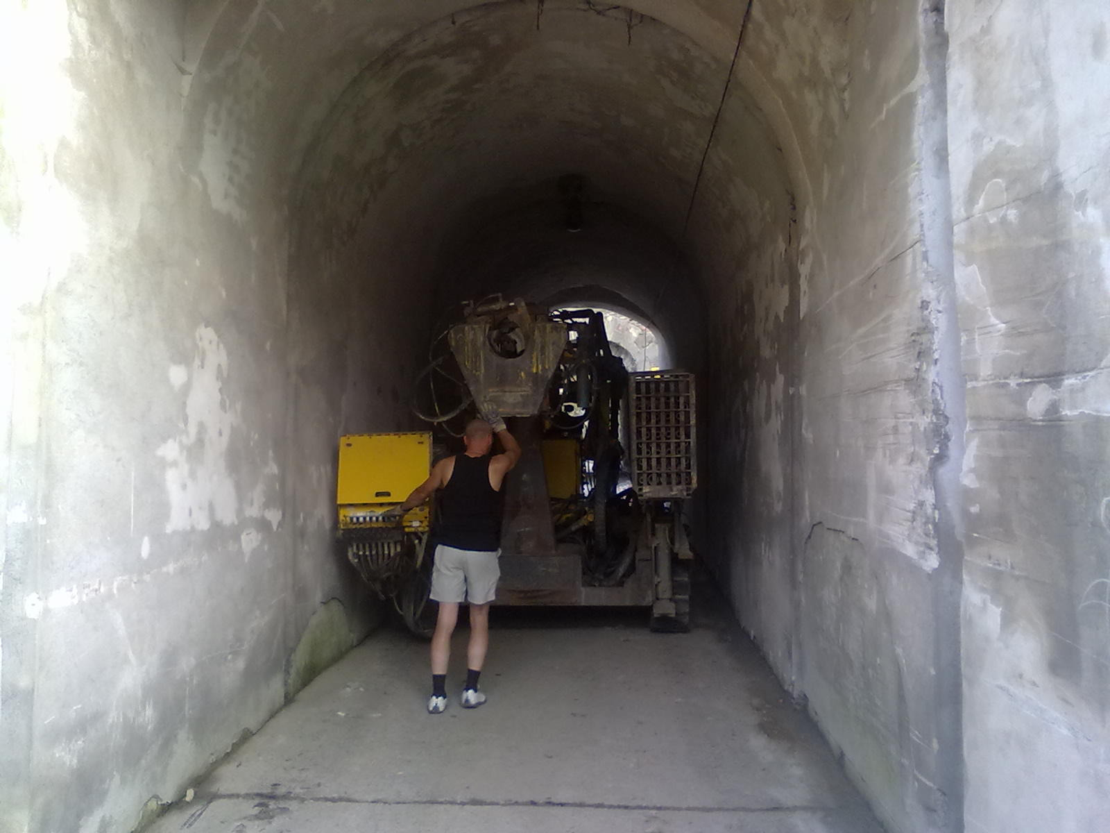 Wnętrze tunelu z maszyną wiertniczą i mężczyzną w podkoszulku, prawdopodobnie podczas prac geologicznych lub wiertniczych. Szare, betonowe ściany tunelu.