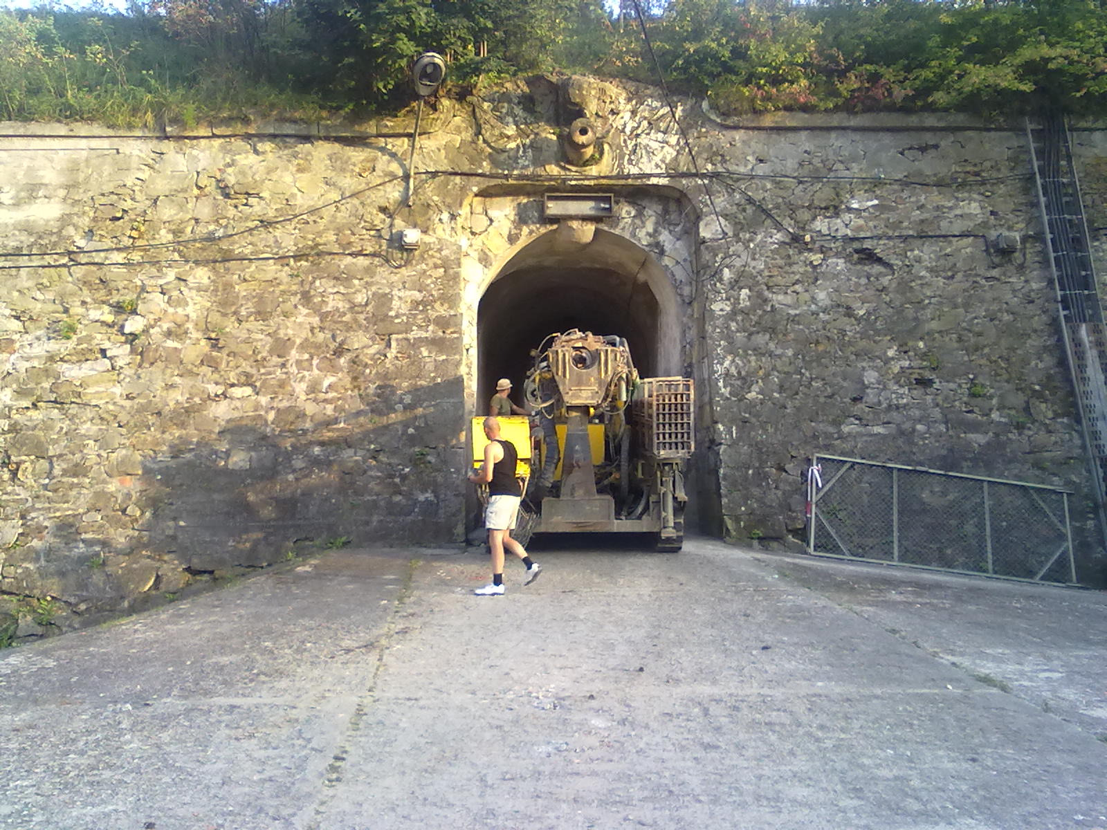 Wnętrze Twierdzy Kłodzkiej z maszyną górniczą i pracownikami. Kamienne mury, tunel, oświetlenie. Osoba w krótkich spodenkach idzie w kierunku maszyny.