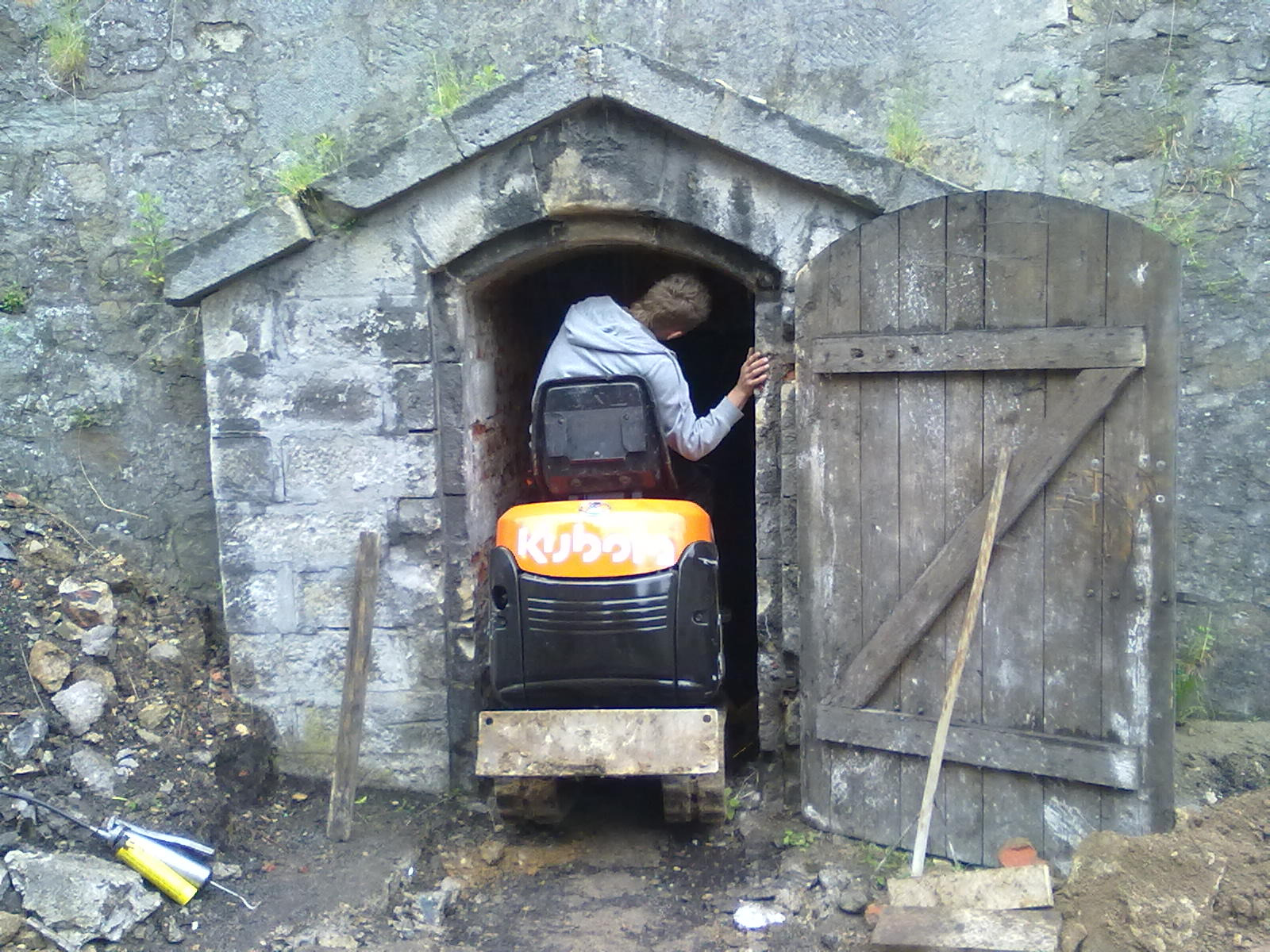 Mini koparka Kubota w kamiennym wejściu z drewnianymi drzwiami, prawdopodobnie w Twierdzy Kłodzko. Osoba wewnątrz. Prace ziemne w toku.