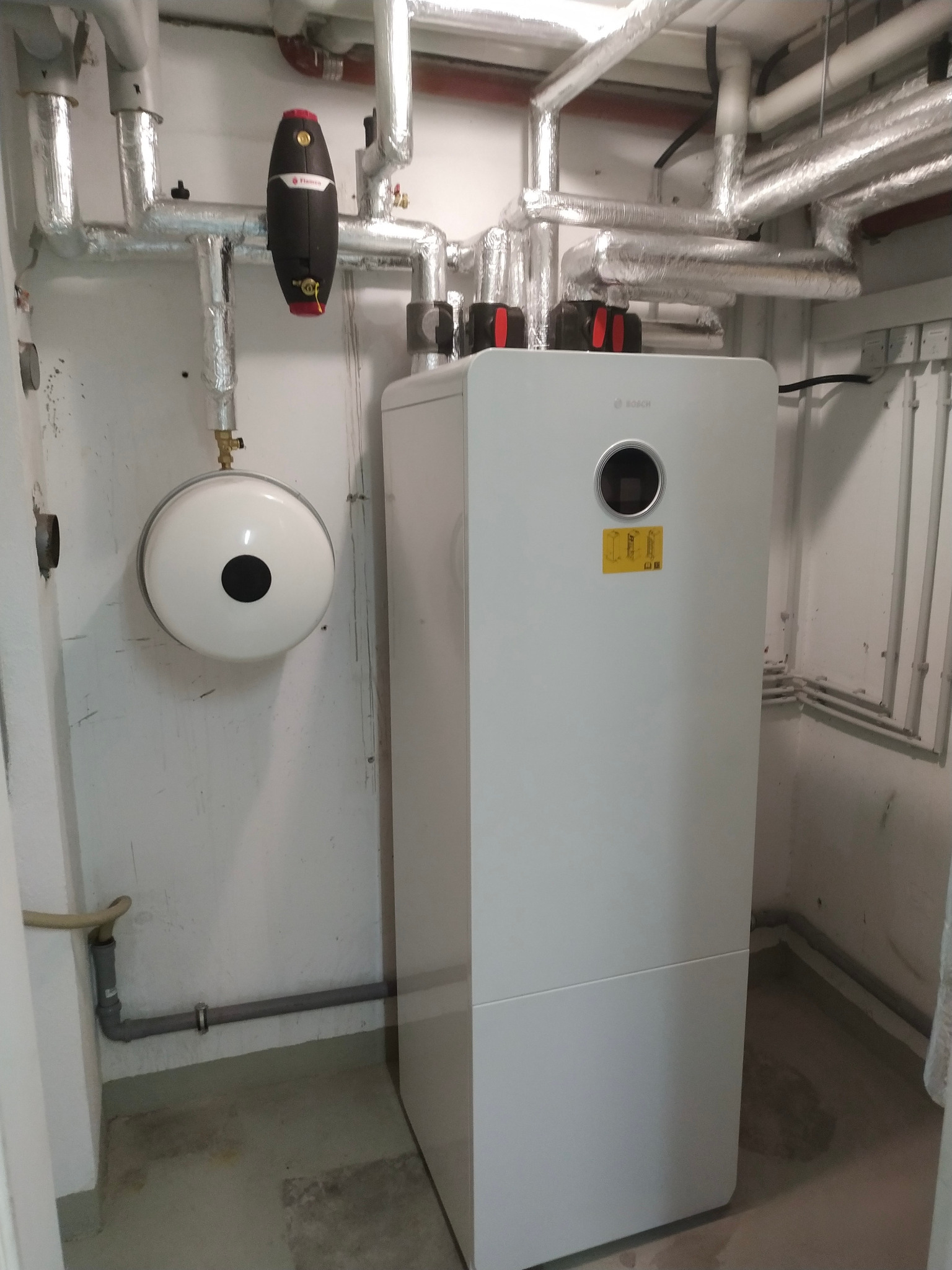 Biała, kompaktowa jednostka w kotłowni z rurami w izolacji. Widoczny zbiornik wyrównawczy i czarny filtr na rurach. Model Bosch z okrągłym panelem sterowania.