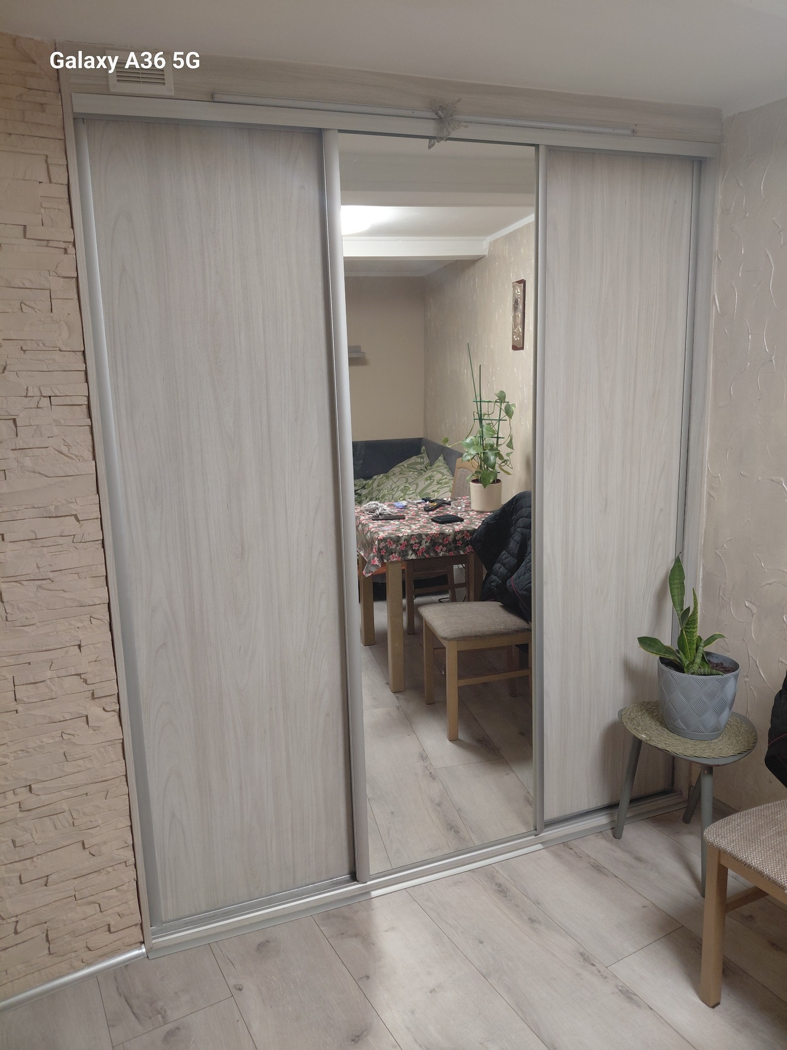 Szafa przesuwna z lustrem w jasnym odcieniu drewna, wkomponowana w pokój z dekoracyjną ścianą z imitacji kamienia i drewnianą podłogą. Widoczne elementy wyposażenia wnętrza.