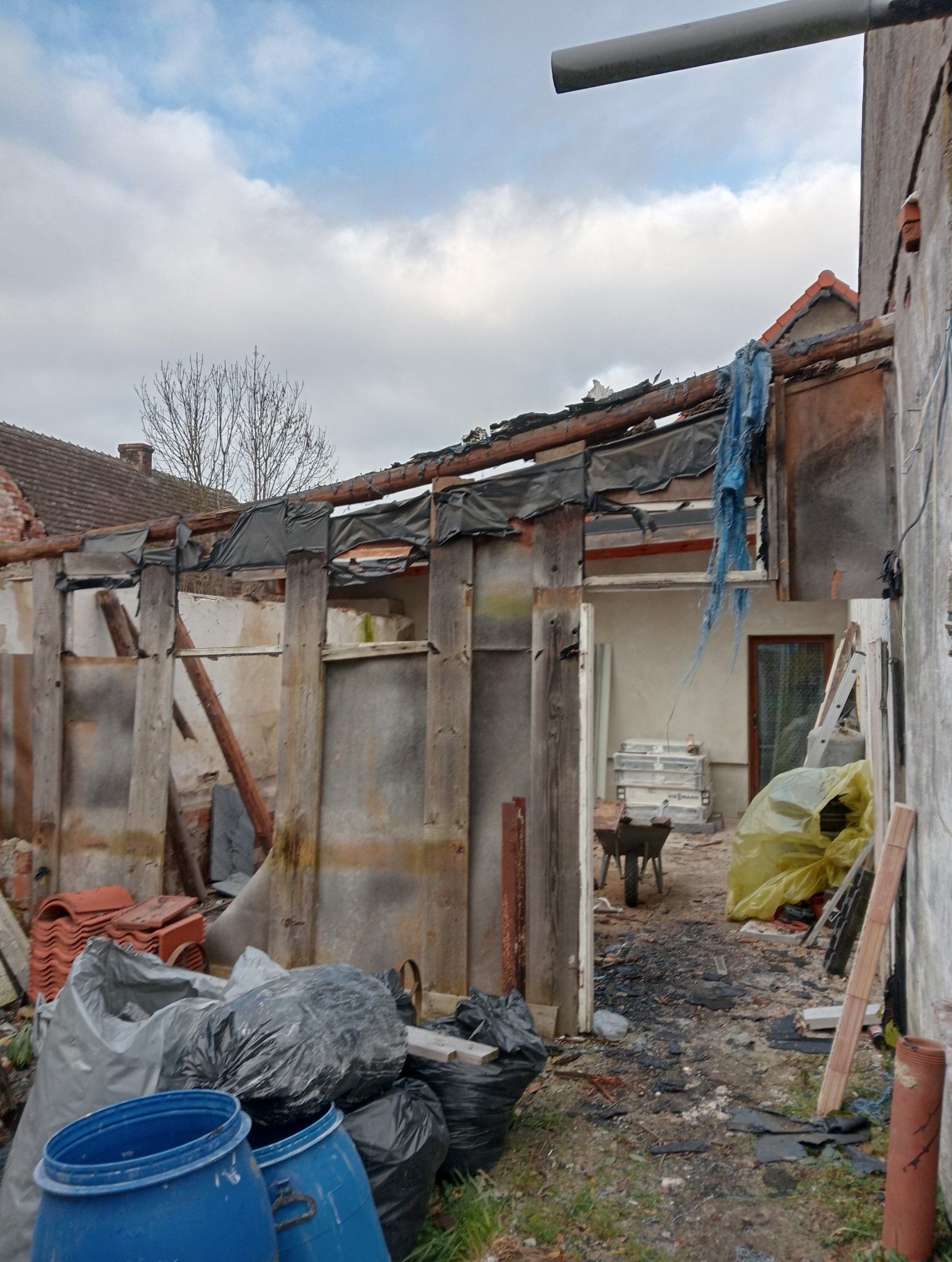 Demontaż konstrukcji dachu z widocznymi elementami drewnianymi, gruzem i workami na odpady. Częściowo zdemontowana konstrukcja, widoczne niebo i fragmenty sąsiednich budynków.