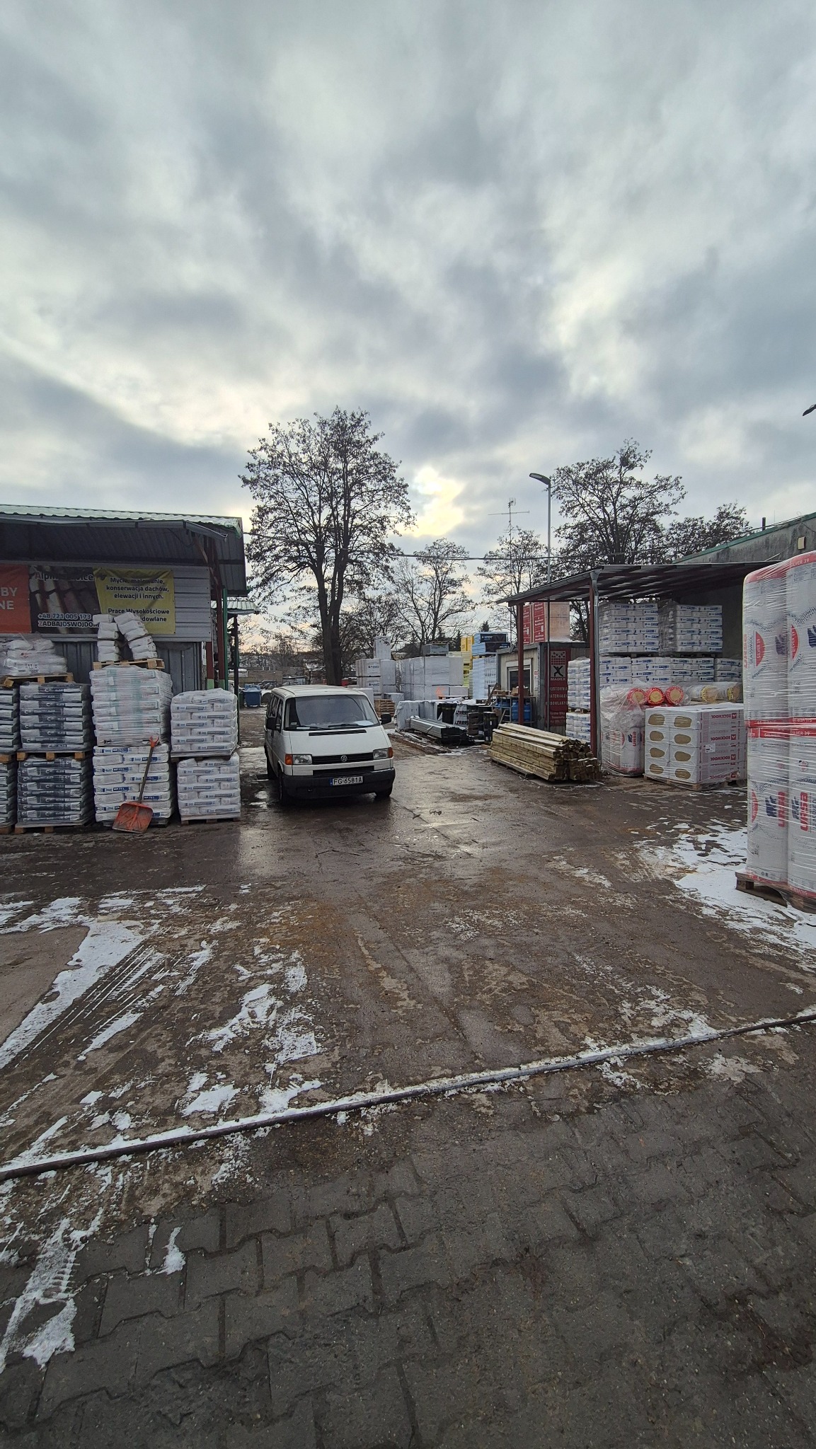 Skład budowlany: palety z materiałami budowlanymi, drewno i samochód dostawczy na placu w pochmurny dzień. Widoczne ślady śniegu.