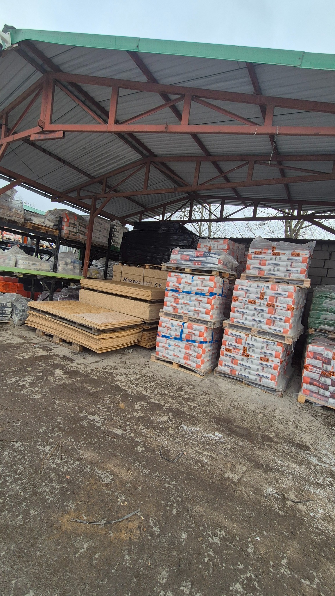 Zadaszony plac składowy z paletami płyt drewnianych i worków z zaprawą, widoczne regały z materiałami budowlanymi w tle, pochmurne niebo.