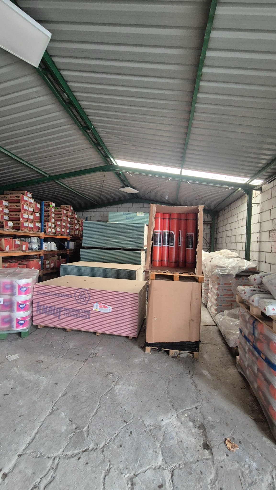 Wnętrze magazynu budowlanego: płyty gipsowo-kartonowe Knauf, materiały izolacyjne w rolkach, worki z zaprawą, regały z produktami. Widok na blaszany dach i betonową posadzkę.