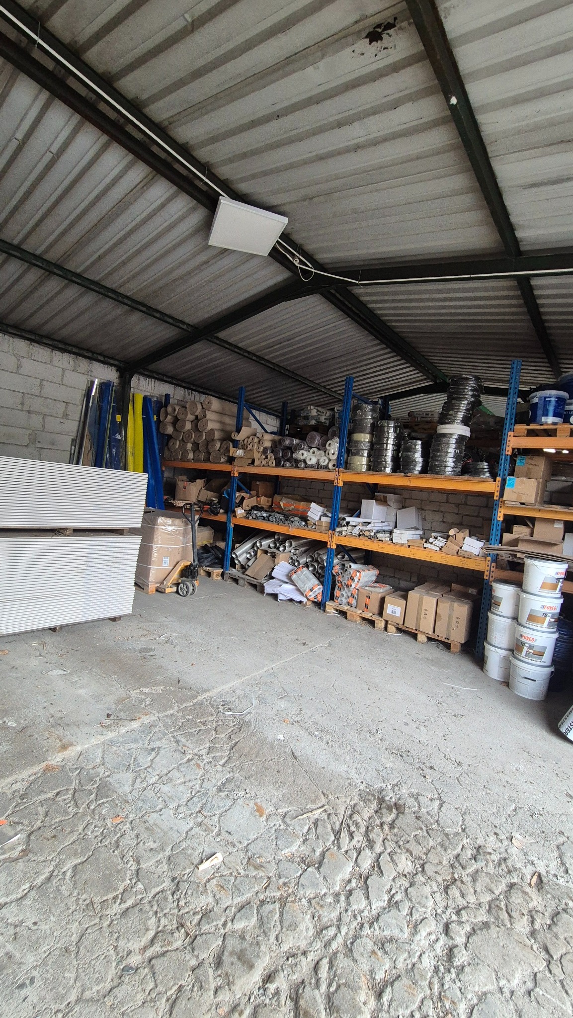 Magazyn z regałami pełnymi materiałów budowlanych, w tym płyty gipsowe, farby i inne akcesoria. Widoczny wózek paletowy.