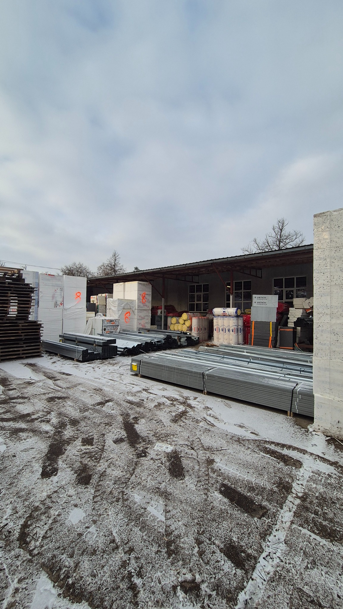 Zimowy widok na skład z materiałami budowlanymi: blachy, wełna i palety. Ślady opon na śniegu prowadzą do magazynu z oknami.
