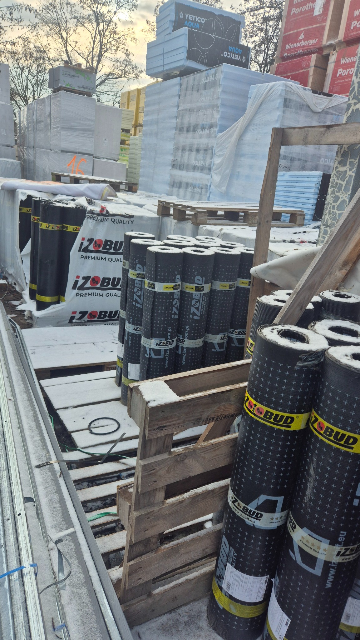 Składowisko materiałów budowlanych zimą: rolki papy bitumicznej Izobud, płyty izolacyjne Yetico i Wienerberger Porotherm na paletach, pokryte śniegiem.