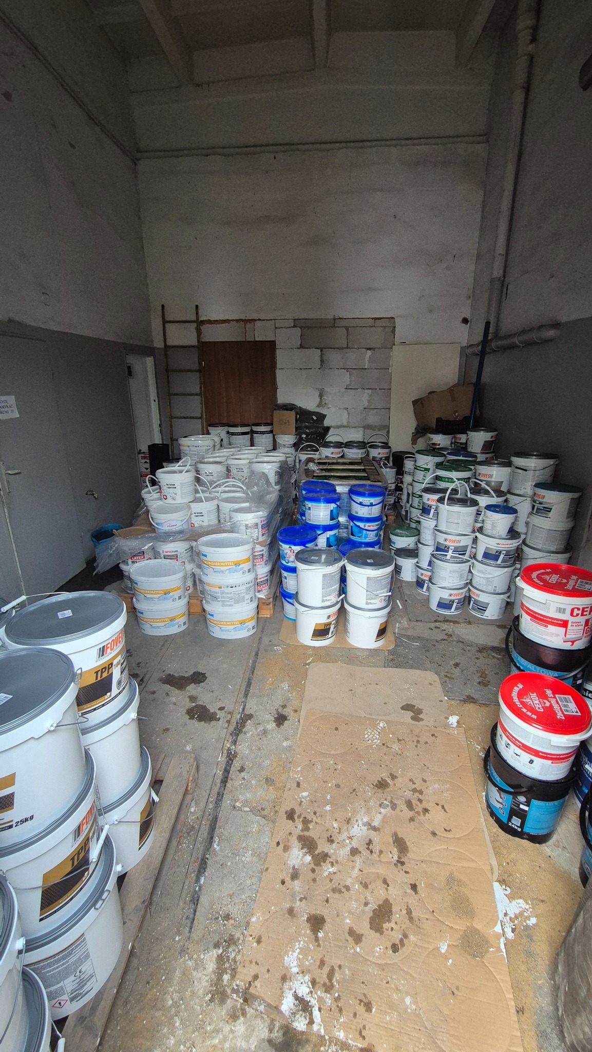 Magazyn z zapasem farb w wiadrach różnych marek i kolorów, ustawionych na podłodze wyłożonej kartonem. Widoczne ślady użytkowania i zabrudzenia.