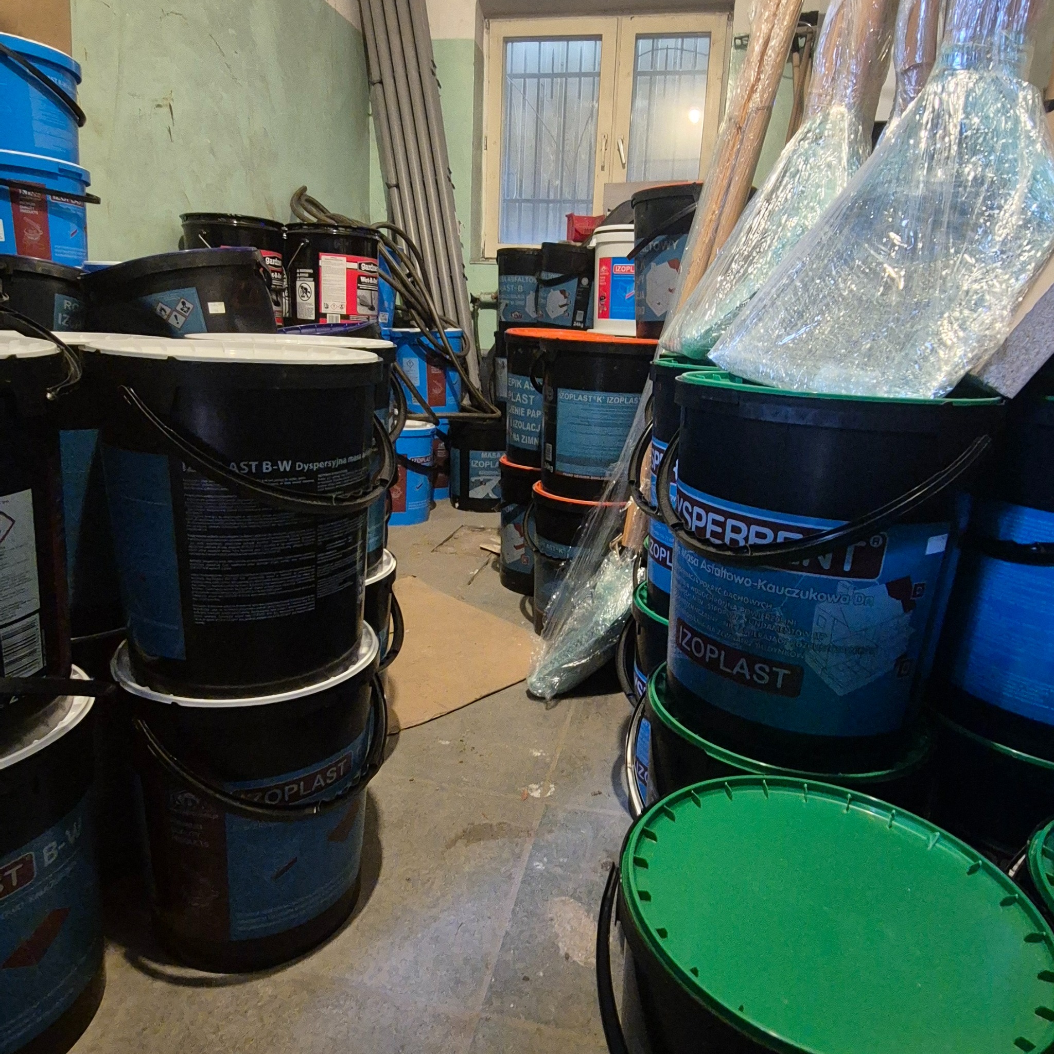 Magazyn z wiadrami masy asfaltowo-kauczukowej Izoplast w kolorach niebieskim, czarnym i zielonym. W tle drabina i okno z kratami.