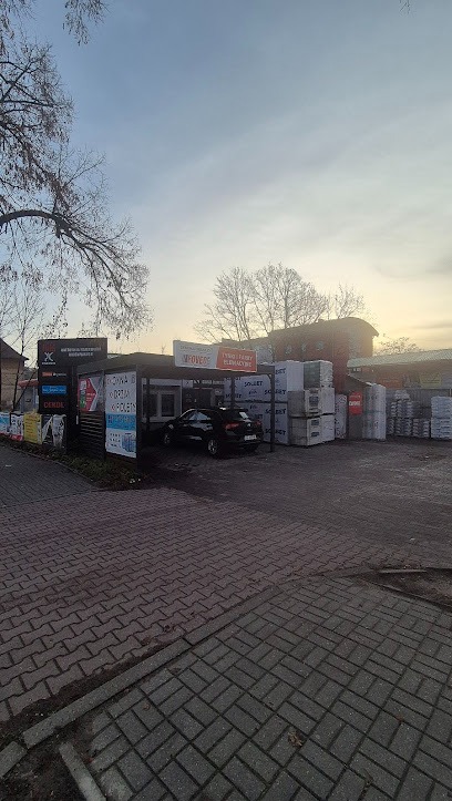 Widok na skład z materiałami budowlanymi i zaparkowanym autem osobowym. Na pierwszym planie kostka brukowa. Widoczne reklamy producentów.