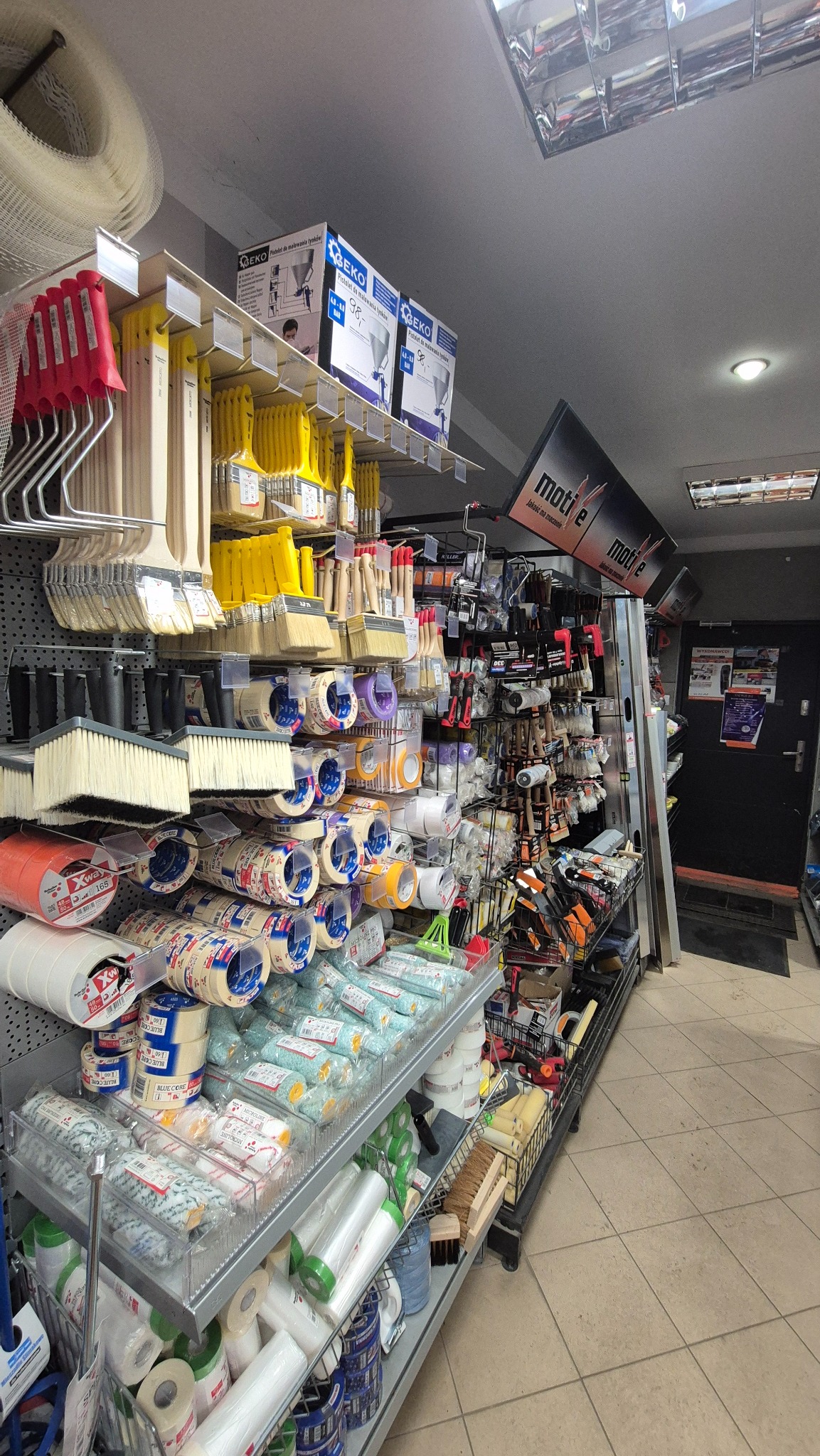 Półki w sklepie z artykułami malarskimi: pędzle, wałki, taśmy malarskie i inne akcesoria. Widok z perspektywy klienta, oświetlenie sufitowe.