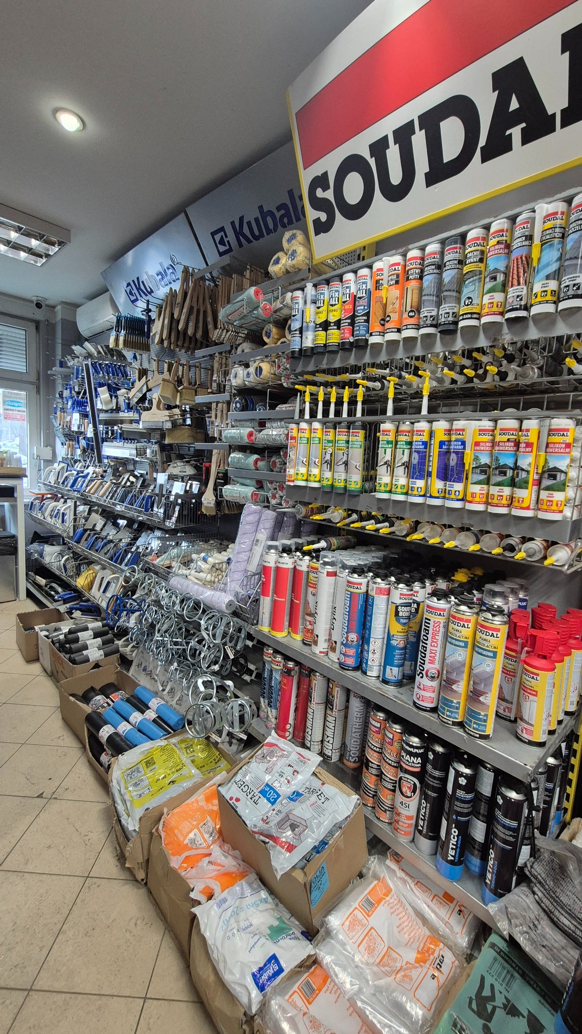 Półki w sklepie budowlanym wypełnione różnorodnymi materiałami: piany montażowe, silikony, pędzle, wałki, narzędzia malarskie i akcesoria. Widoczne logo Soudal i Kubala.