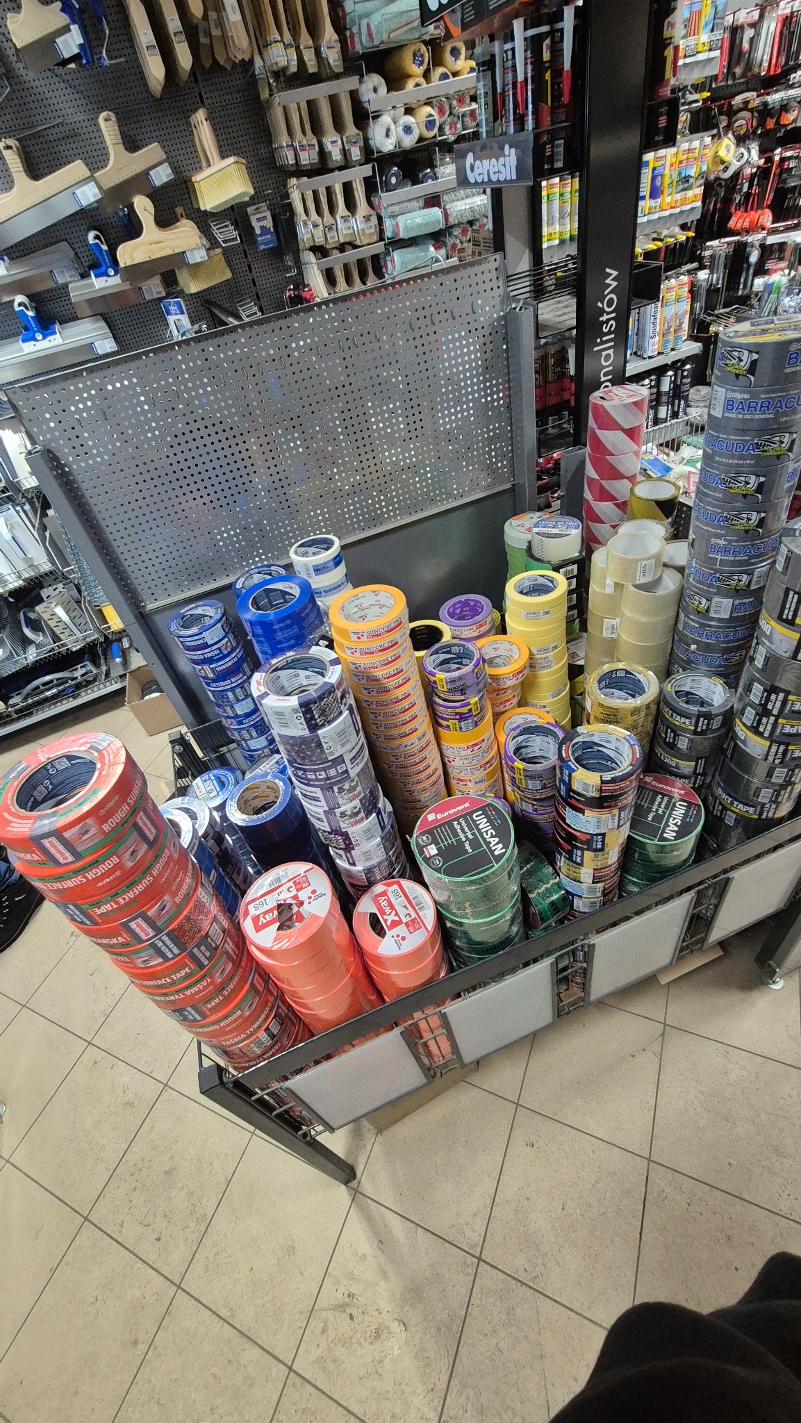 Ekspozycja różnorodnych taśm klejących w sklepie budowlanym. Widoczne taśmy malarskie, uniwersalne i specjalistyczne, ułożone w stosy na metalowym regale.