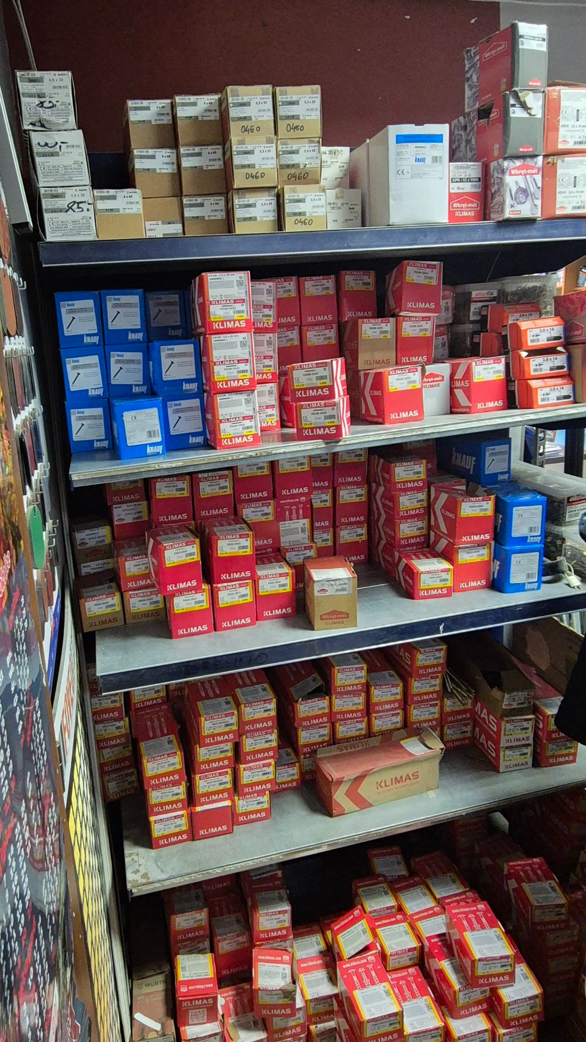 Półka w sklepie z artykułami budowlanymi, wypełniona pudełkami wkrętów i kołków firm Klimas i Knauf. Różne rozmiary i typy, ułożone rzędami. Widoczne etykiety z oznaczeniami.