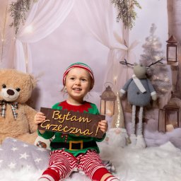 Pogodna fotografia - Uśmiechnięta dziewczynka w stroju elfa trzyma tabliczkę z napisem 'Byłam grzeczna!' na tle świątecznej scenografii z pluszowym misiem i reniferem.