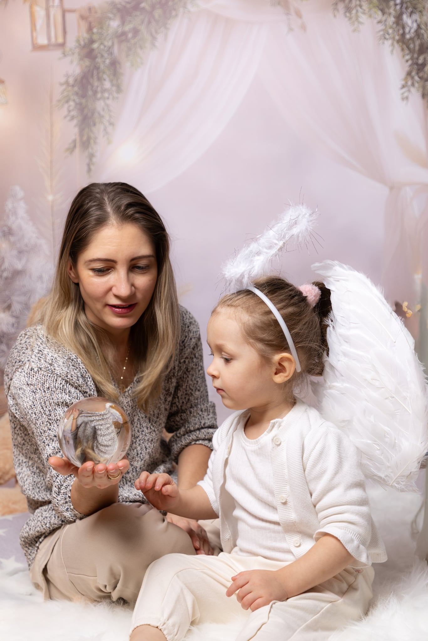 Mama z córką w stroju anioła, siedząca na puszystym dywanie, trzymająca szklaną kulę z odbiciem wnętrza. Delikatne, pastelowe tło z motywem zimowym.