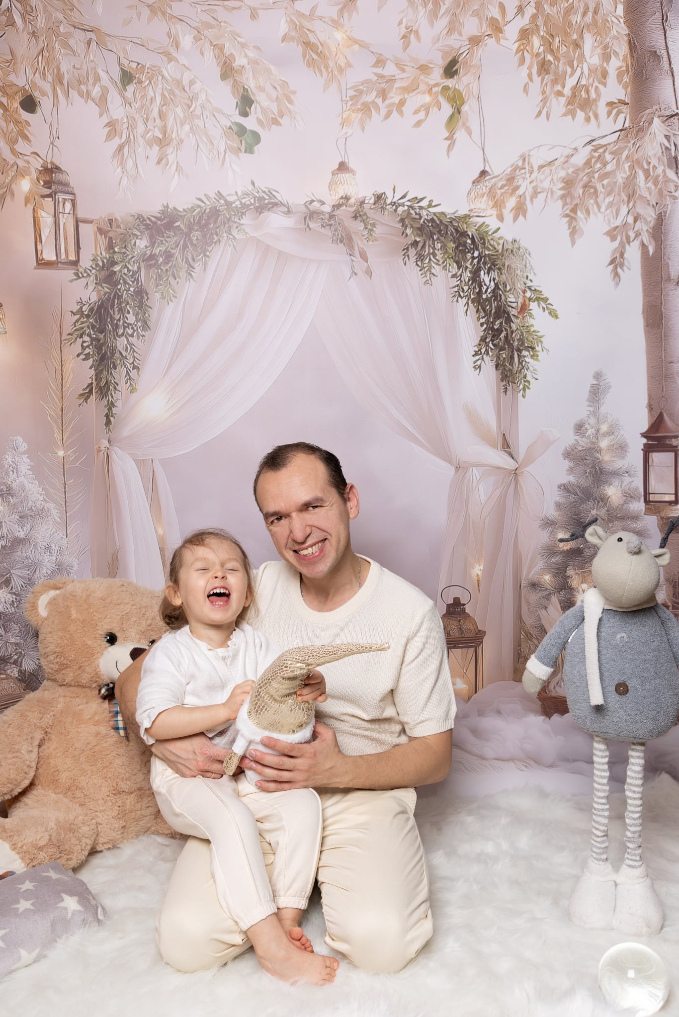 Uśmiechnięty tata z córeczką na kolanach, w świątecznej scenerii z pluszakami i dekoracjami. Dziewczynka śmieje się, trzymając zabawkę.