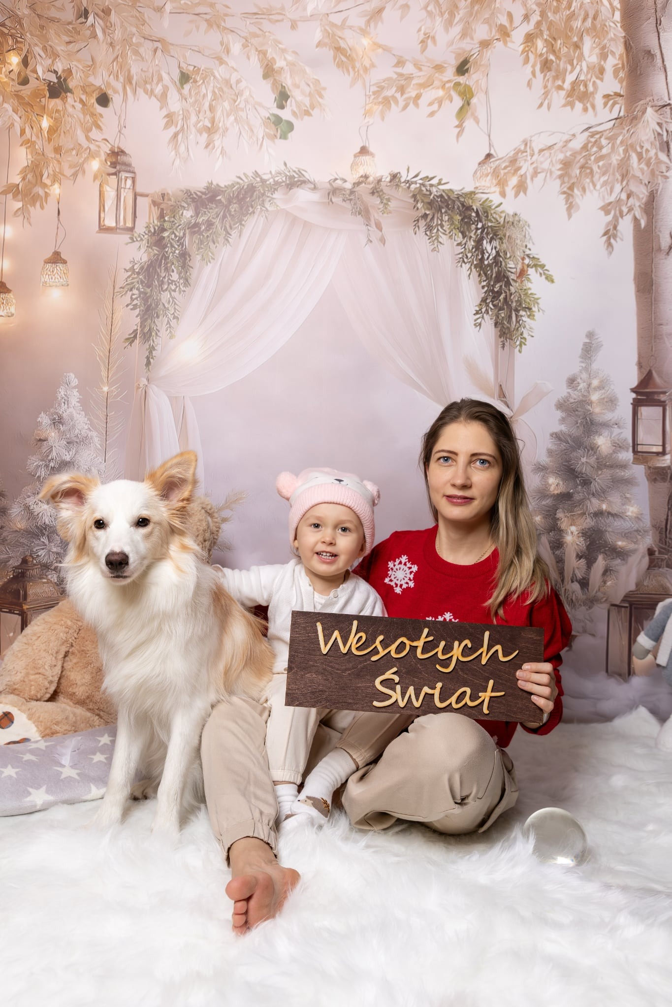 Świąteczna sesja: Mama z dzieckiem i psem trzymają drewniany napis 'Wesołych Świąt' na tle dekoracji z choinkami i lampionami. Dziewczynka w różowej czapce.