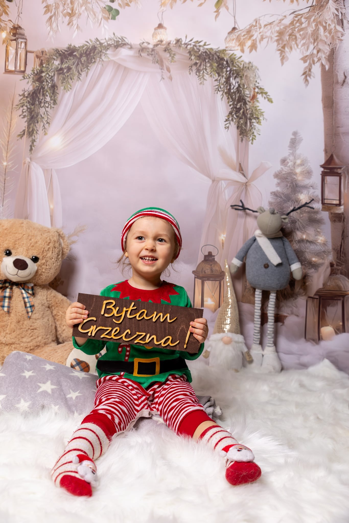 Uśmiechnięta dziewczynka w stroju elfa trzyma tabliczkę z napisem 'Byłam grzeczna!' na tle świątecznej scenografii z pluszowym misiem i reniferem.