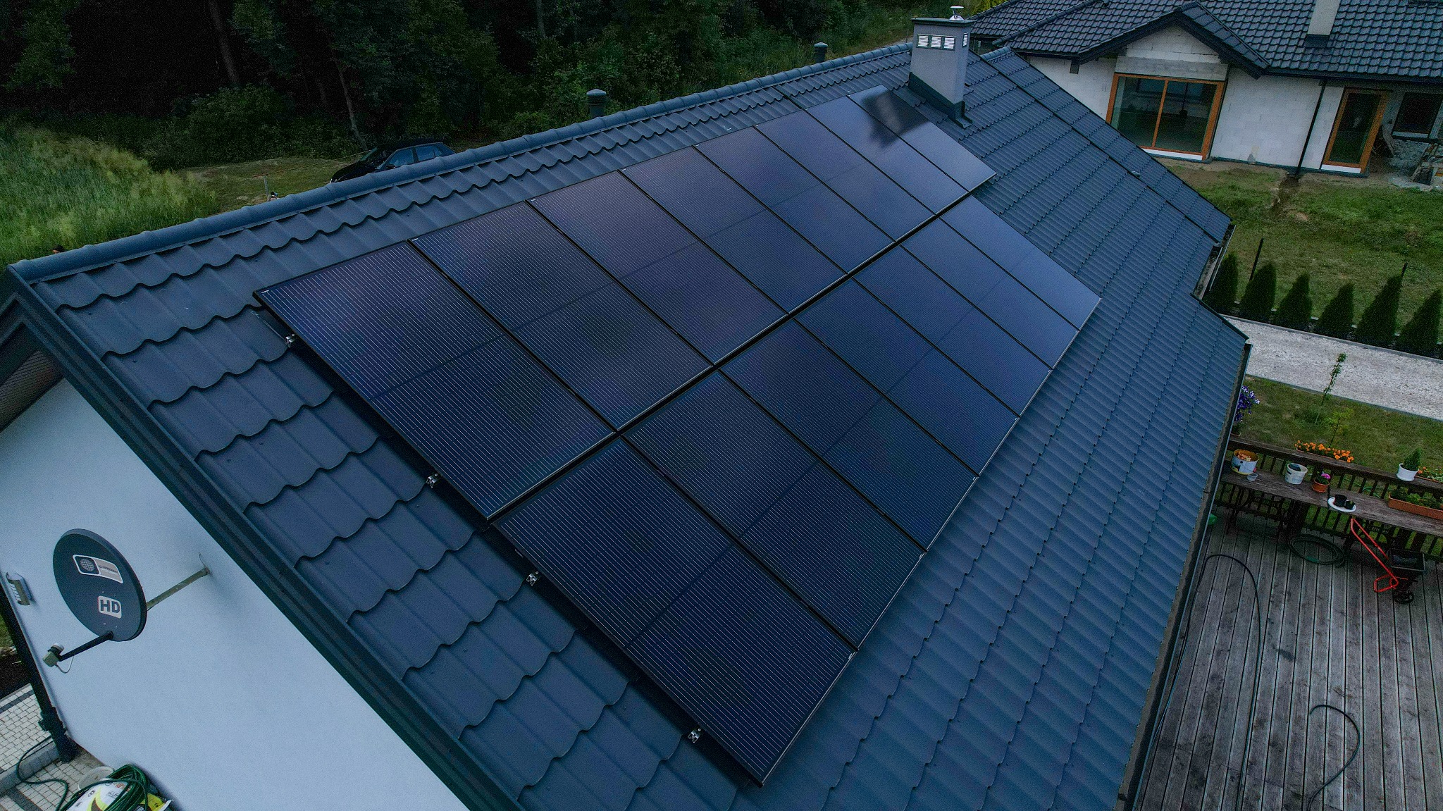 Panele fotowoltaiczne na dachu domu jednorodzinnego, widok z góry. Ciemna dachówka i nowoczesna instalacja PV. Widoczny fragment ogrodu i sąsiedni budynek.