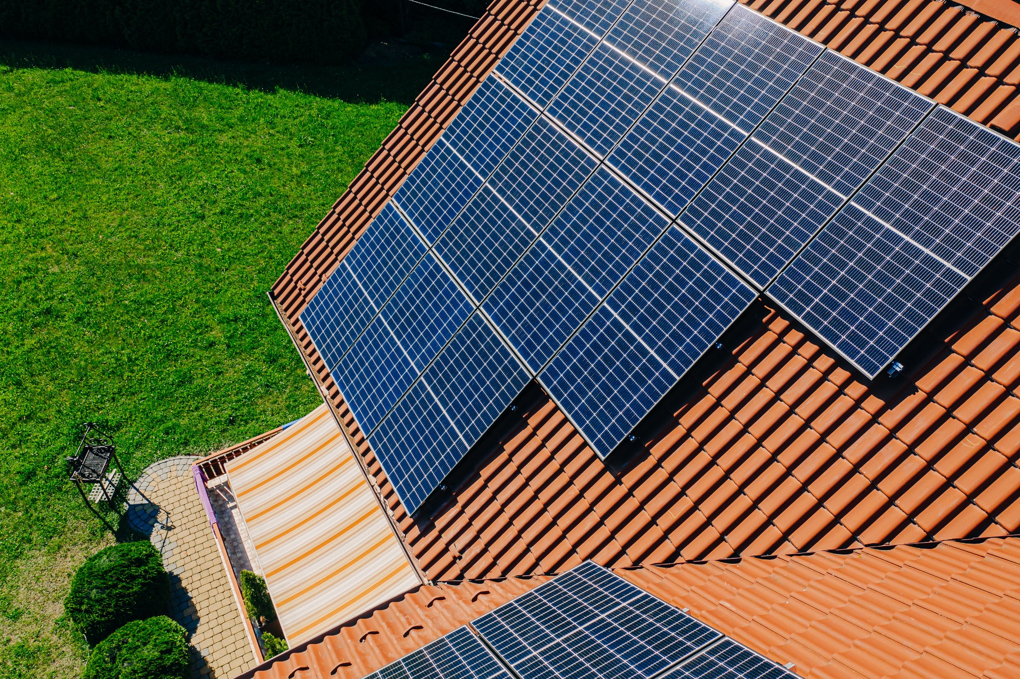 Widok z góry na panele fotowoltaiczne na czerwonym dachu domu, obok zielony trawnik i markiza w paski. Część paneli jest ucięta w kadrze.