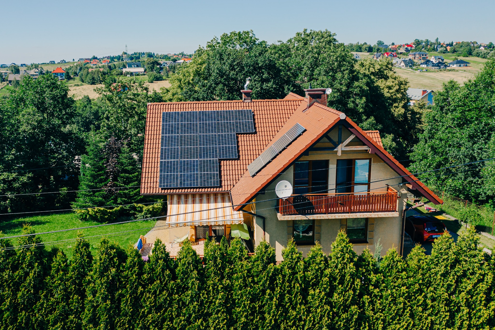 Dom z panelami słonecznymi na dachu z czerwonej dachówki, widok z góry, otoczony zielenią. W tle zabudowa wiejska i drzewa.