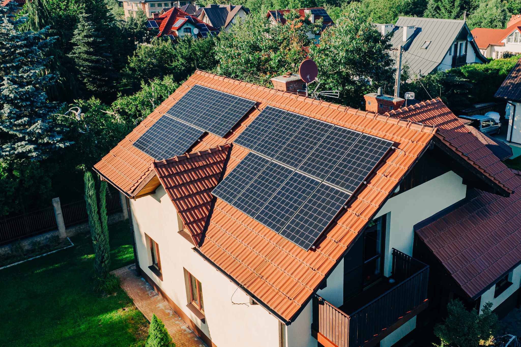 Dom jednorodzinny z panelami fotowoltaicznymi na dachu z czerwonej dachówki, widok z góry, antena satelitarna na kominie, zielone otoczenie.