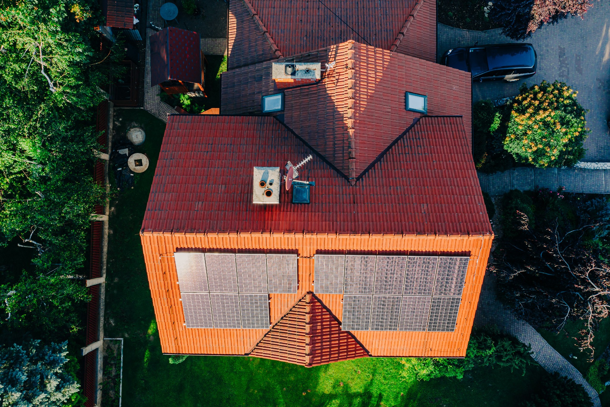 Widok z drona na dom z czerwoną dachówką i panelami słonecznymi. Obok domu zaparkowany samochód i zadbany ogród z drzewami.