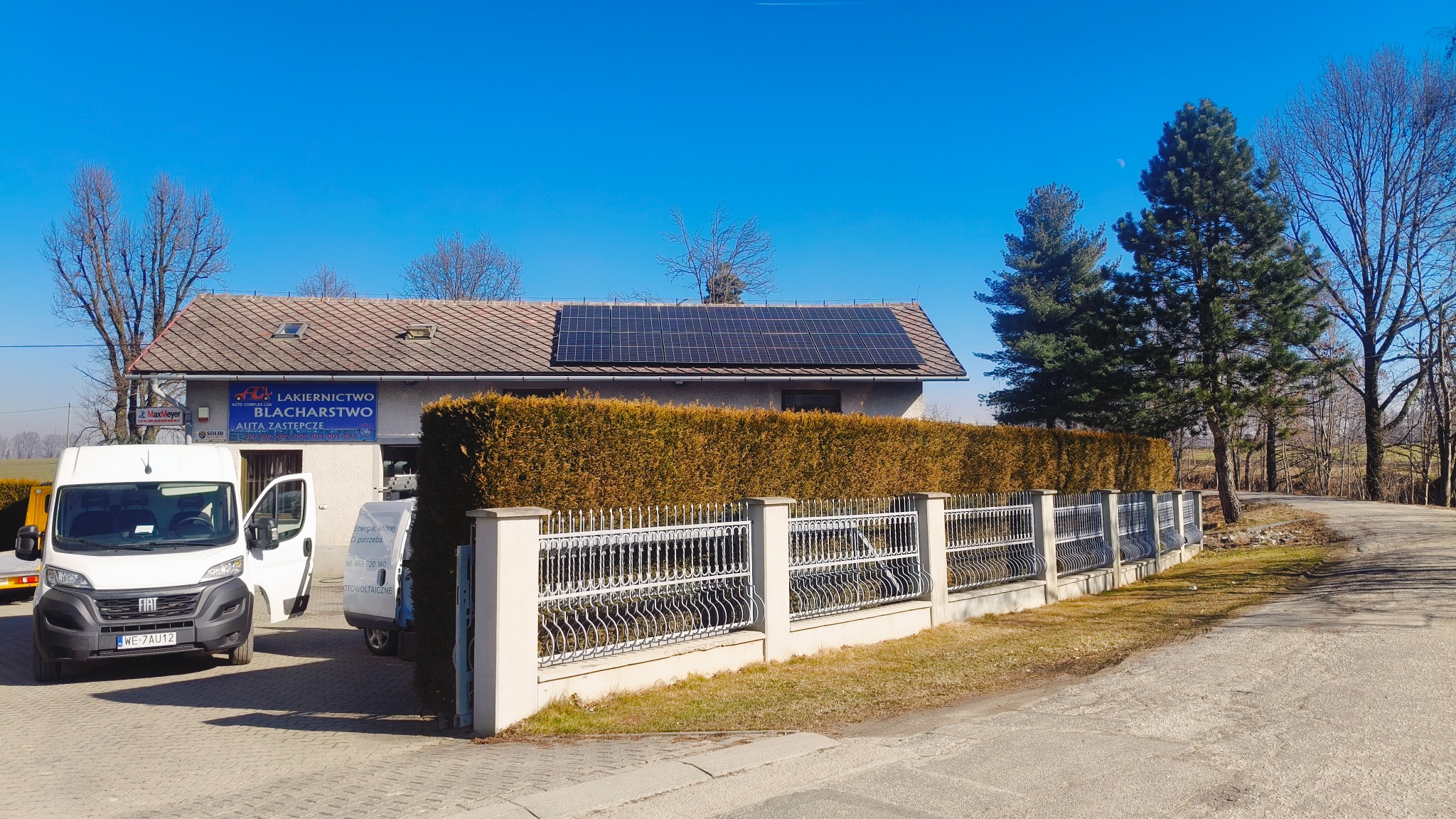 Budynek z panelami słonecznymi na dachu, widoczny zza ogrodzenia i żywopłotu. Na pierwszym planie zaparkowane busy. Błękitne niebo w tle.