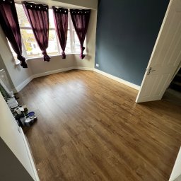 Krzysztof Sowinski - Pokój po remoncie: drewniana podłoga, granatowa ściana, okno z fioletowymi zasłonami. Widoczne puszki z farbą, sugerujące świeżo zakończone prace malarskie.