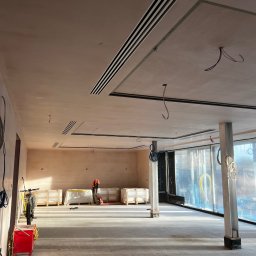 Krzysztof Sowinski - Wnętrze w trakcie wykończenia: gładzie na ścianach, kratki wentylacyjne w suficie, folia ochronna na oknach. Widoczny pracownik w oddali i materiały budowlane.