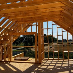 Krzysztof Sowinski - Drewniana konstrukcja dachu w trakcie budowy, widok z wnętrza. Elementy więźby dachowej z jasnego drewna na tle błękitnego nieba i krajobrazu z polami.