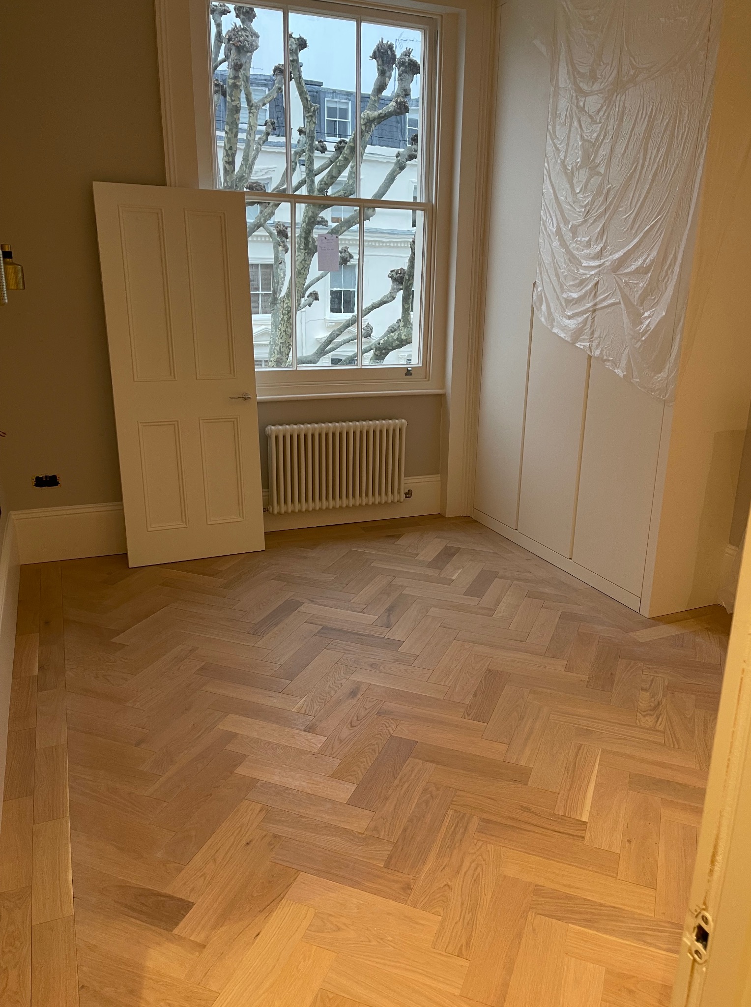 Jasny parkiet ułożony w jodełkę francuską w pokoju z oknem i grzejnikiem. Białe drzwi oparte o ścianę, zabudowana szafa z folią ochronną. Widok na drzewa za oknem.