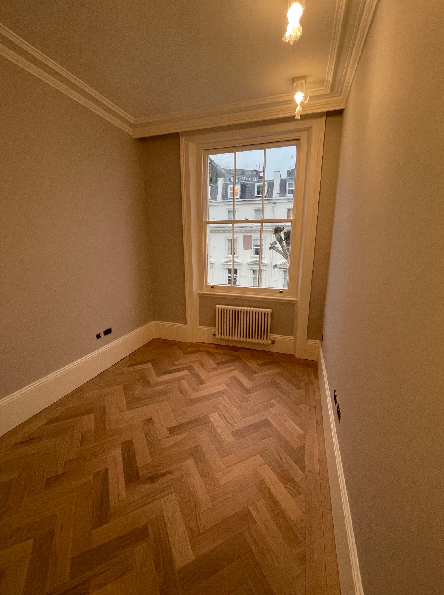 Pokój po remoncie: parkiet w jodełkę, świeżo pomalowane ściany w odcieniach beżu, białe listwy przypodłogowe i okienne. Widok na budynek przez okno.