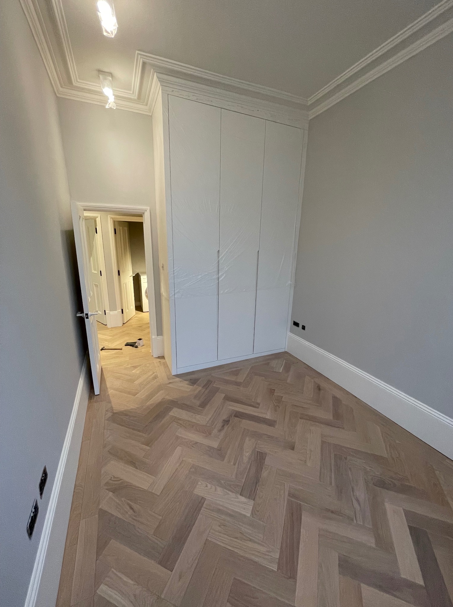 Wnętrze pokoju po remoncie: parkiet w jodełkę, biała szafa wnękowa zabezpieczona folią, listwy przypodłogowe i sufitowe, otwarte drzwi do kolejnego pomieszczenia.